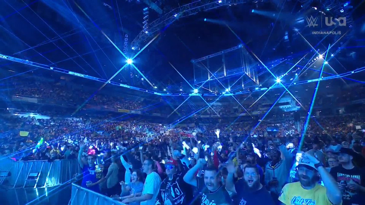 A massive WWE crowd cheers enthusiastically inside a packed arena illuminated by crisscrossing blue laser beams, with the broadcast graphic in the upper right reading "WWE USA INDIANAPOLIS," setting the stage for the cage match entrance.