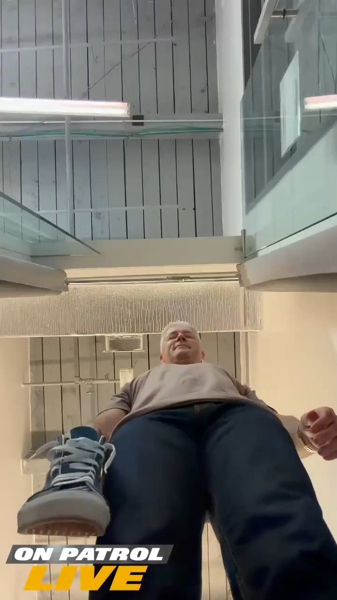 A low-angle shot from a TV show 'ON PATROL LIVE' showing a heavyset man in a tan shirt and dark pants with one leg lifted, viewed from below on a staircase landing with glass railings.