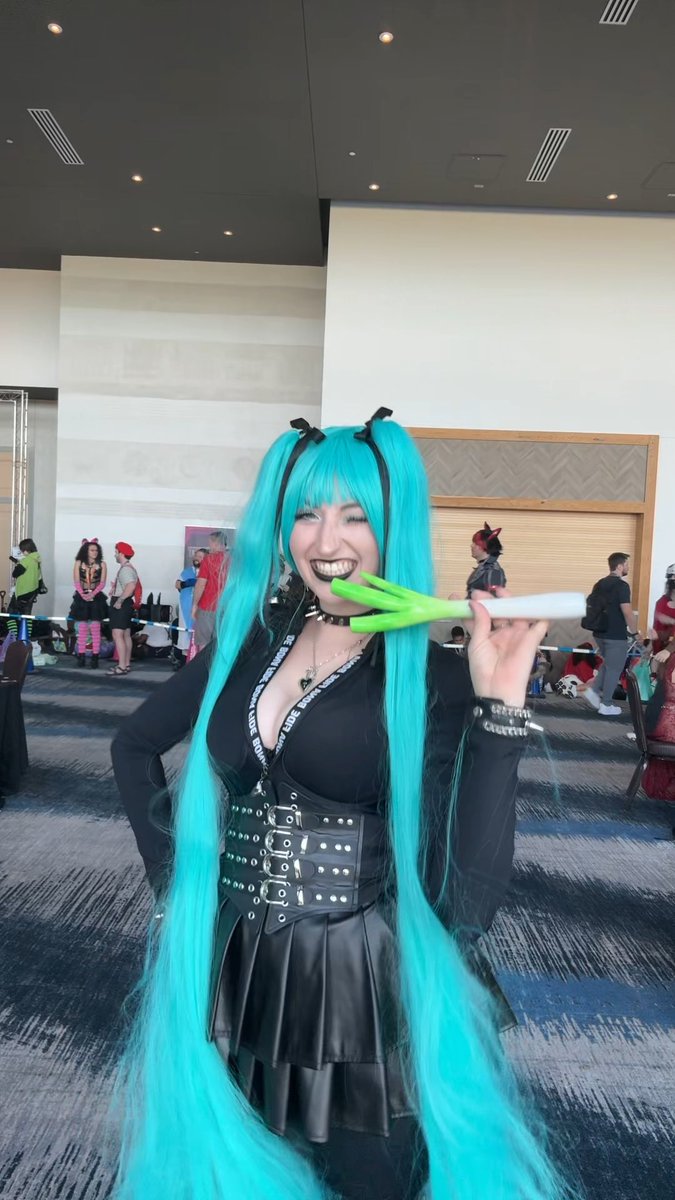A cosplayer with long teal twin-tails winks playfully while holding a prop with green blades, dressed in a black gothic-style outfit at a convention.