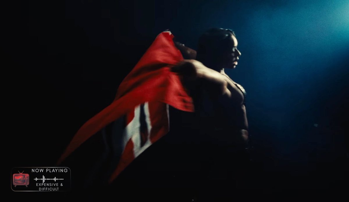 A person stands in dramatic low lighting, holding a red flag or cape. Text in the corner reads 'NOW PLAYING EXPENSIVE & DIFFICULT'.