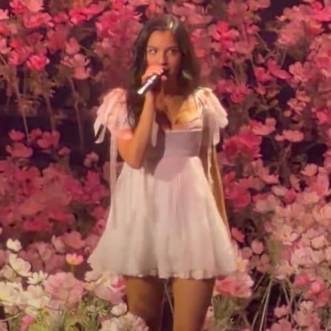 A woman in a white dress performing on stage with a microphone against a backdrop of pink flowers.