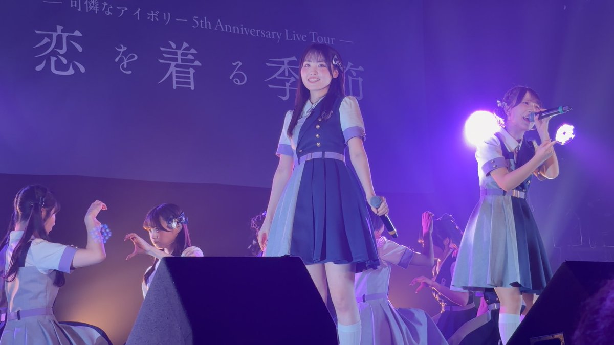 Young female performers in matching blue and white sailor-style outfits sing and dance on stage at a concert, with text on the screen reading '5th Anniversary Live Tour' in Japanese.