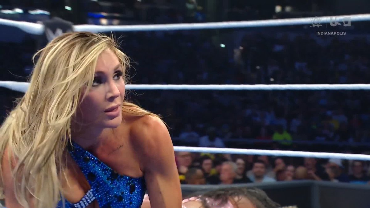 A blonde female wrestler in a blue sparkly outfit looks intently across the ring at a WWE event in Indianapolis, as captured by a USA Network broadcast, with the text noting Charlotte Flair ("The Queen") executing a "Double Moonsault" move.