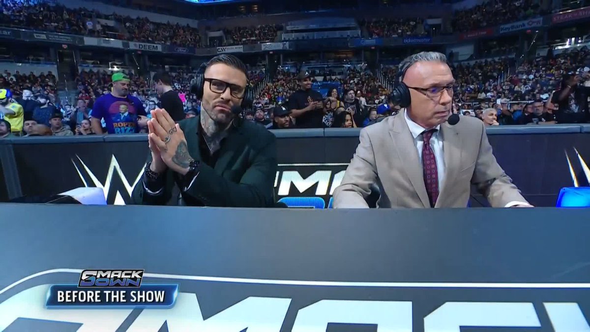 Two WWE commentators sit at the announce desk during a "SmackDown Before the Show" segment, with a crowd visible behind them and a fan wearing a "Ropes Rizz" shirt in the stands. The image appears to be from a WWE broadcast, though the commentators pictured don't match the Randy Orton interview mentioned in the caption.