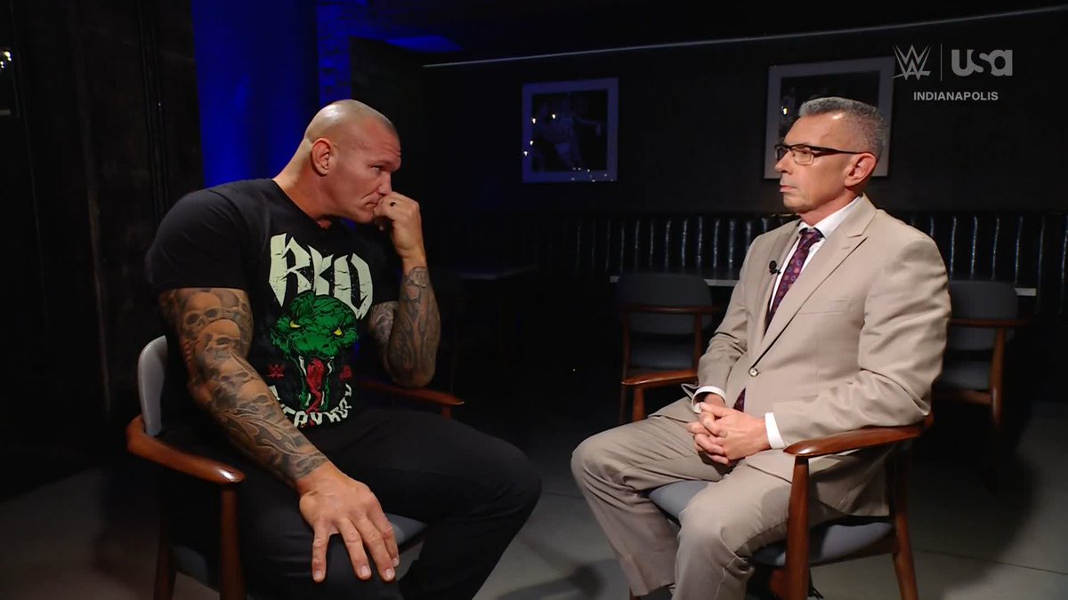 A muscular, tattooed wrestler in a black "RKO" graphic t-shirt sits hunched forward with his hand to his mouth, looking contemplative as he faces an interviewer in a tan suit and glasses. The WWE/USA Network logo with "INDIANAPOLIS" appears in the upper right corner, fitting the post's note about an awkward interview moment where the wrestler seems to be carefully choosing his words.