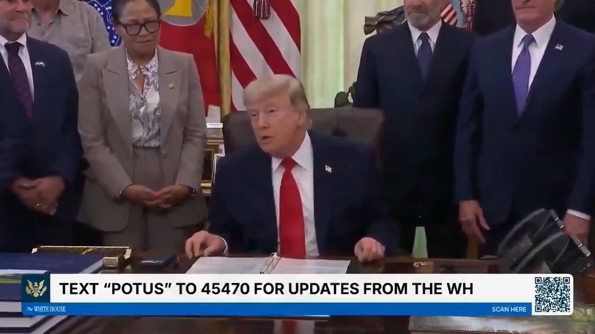 Donald Trump sits at the Resolute Desk in the Oval Office surrounded by officials, with a chyron reading 'TEXT "POTUS" TO 45470 FOR UPDATES FROM THE WH' alongside 'The WHITE HOUSE' logo.