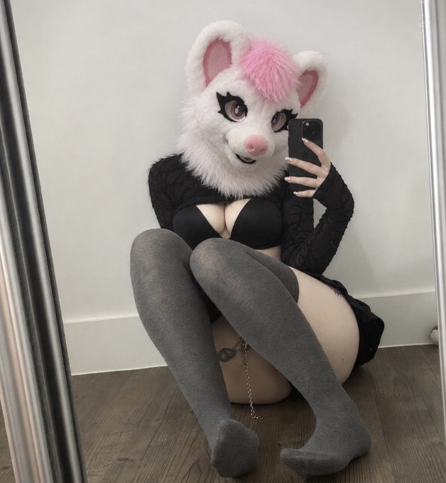 A person wearing a fluffy white and pink animal mask, a black top, thigh-high gray socks, and a garter belt sits on the floor taking a mirror selfie.