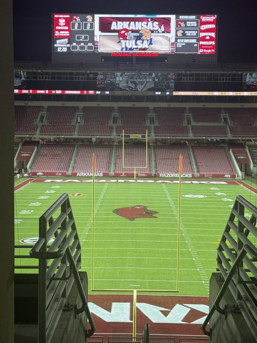 A nighttime view from inside a stadium shows an empty football field with an Arkansas Razorbacks logo at midfield, and a scoreboard displaying 'ARKANSAS vs TULSA'.