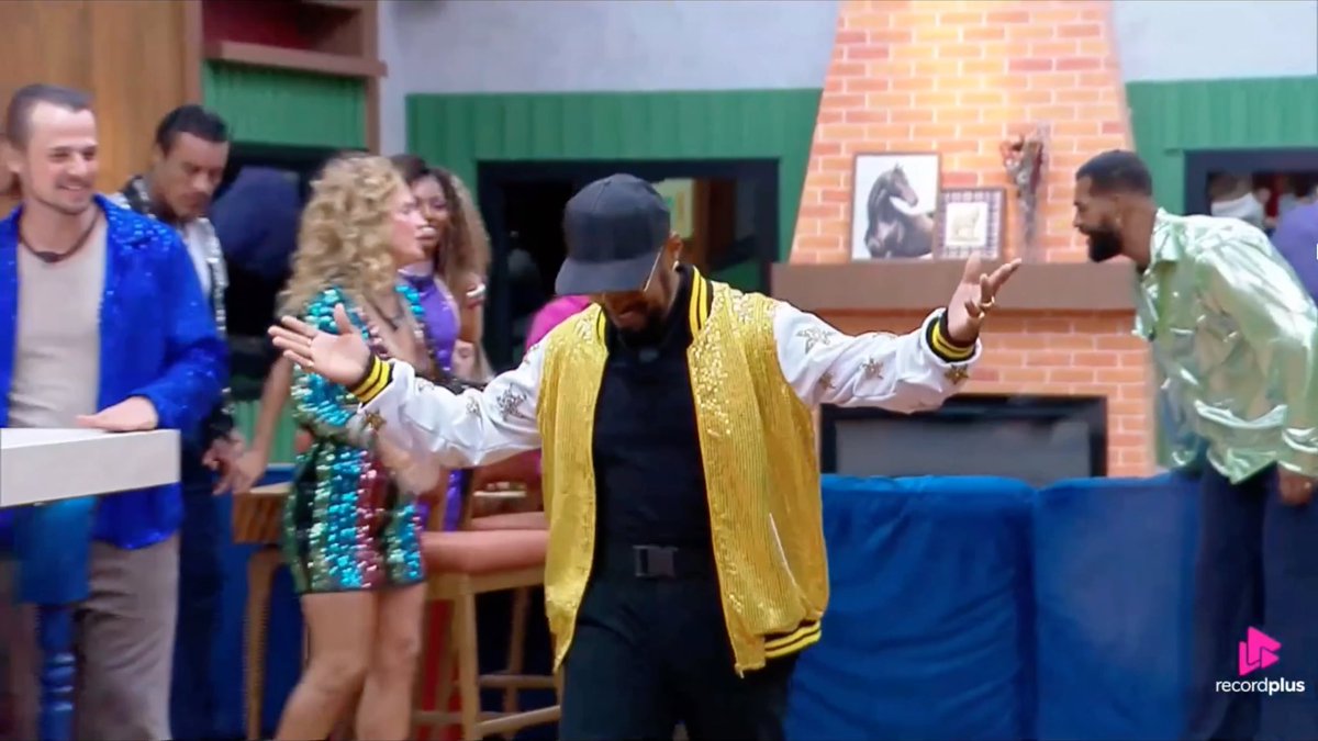 A screenshot from a TV show or video featuring a man in a sparkly yellow jacket and black cap dancing or gesturing with his arms out, surrounded by other people in a colorful, eclectic room setting; a 'recordplus' watermark is in the bottom right corner.