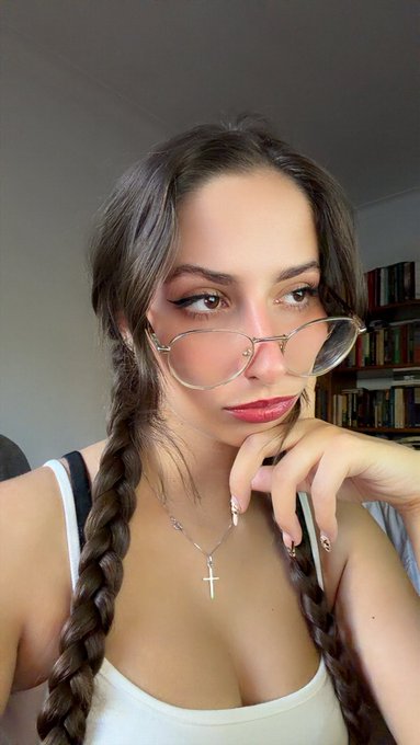 A selfie of a young woman with long brown braided hair, wearing pink-tinted glasses and a silver cross necklace, with a bookshelf visible in the background.