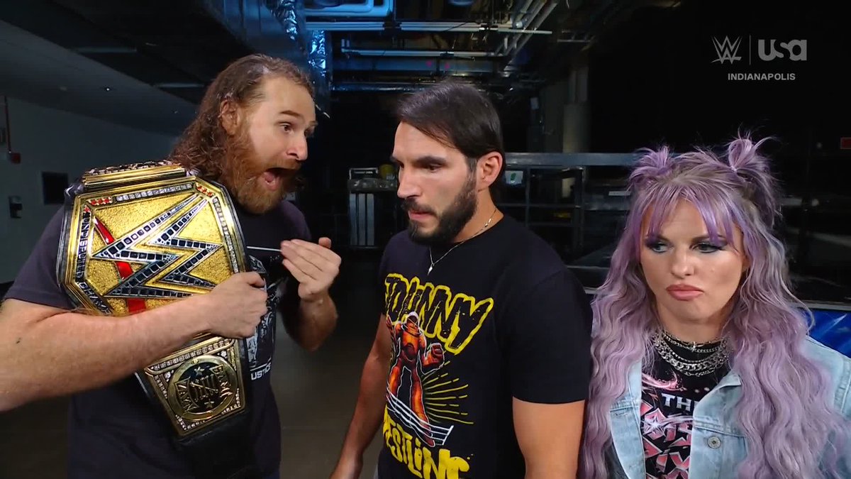 A long-haired bearded wrestler holds up an NXT Championship belt while enthusiastically addressing two stern-looking opponents—a bearded man in a yellow graphic tee and a woman with lavender pigtails—backstage in Indianapolis, capturing the post's "Ride or Die" energy of an unflinching championship confrontation.