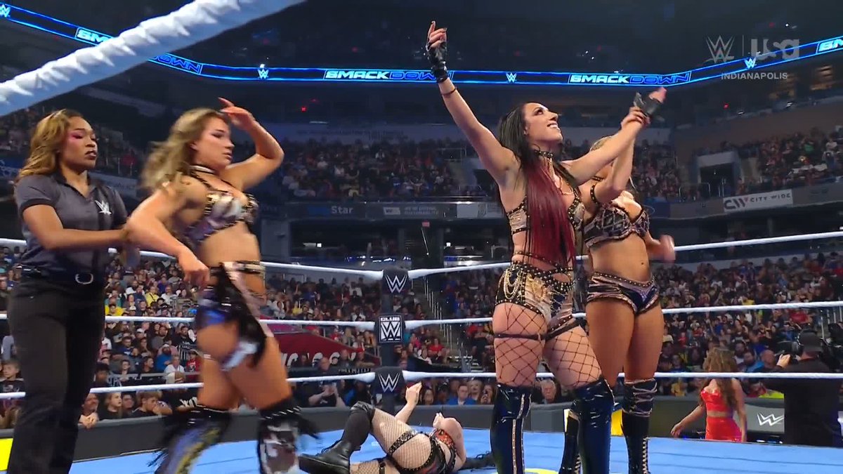 The image shows a WWE SmackDown match in Indianapolis where two wrestlers, Fallon Henley and Lainey Reid, stand victorious in the ring with their arms raised in celebration, while an opponent lies defeated on the mat and a referee observes nearby, capturing the moment Fatal Influence secured their win.