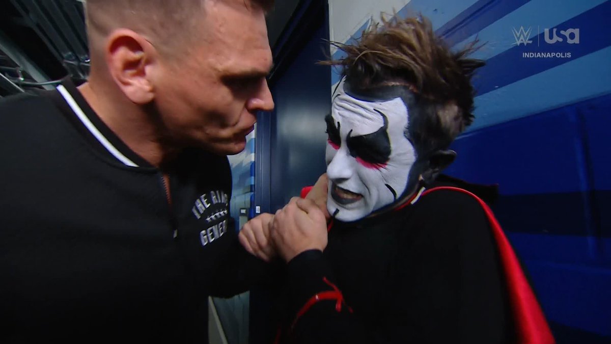 Two men are face-to-face in a tense confrontation, with one grabbing the other by the collar. The man on the left, wearing a black shirt with "THE RING GENE..." visible, is locked in a heated stare with the man on the right, who sports elaborate black, white, and red face paint with spiky hair. The WWE on USA logo and "INDIANAPOLIS" text appear in the upper right, capturing what looks like an altercation between Gunther and Danhausen at a wrestling event.