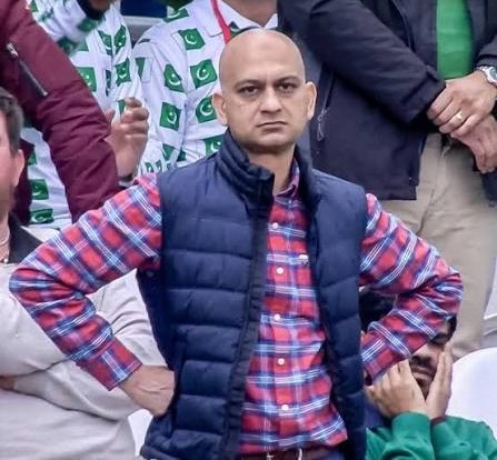A photo of a bald man wearing a blue puffer vest over a red plaid shirt, standing with his hands on his hips at what appears to be a sports event with people in the background.