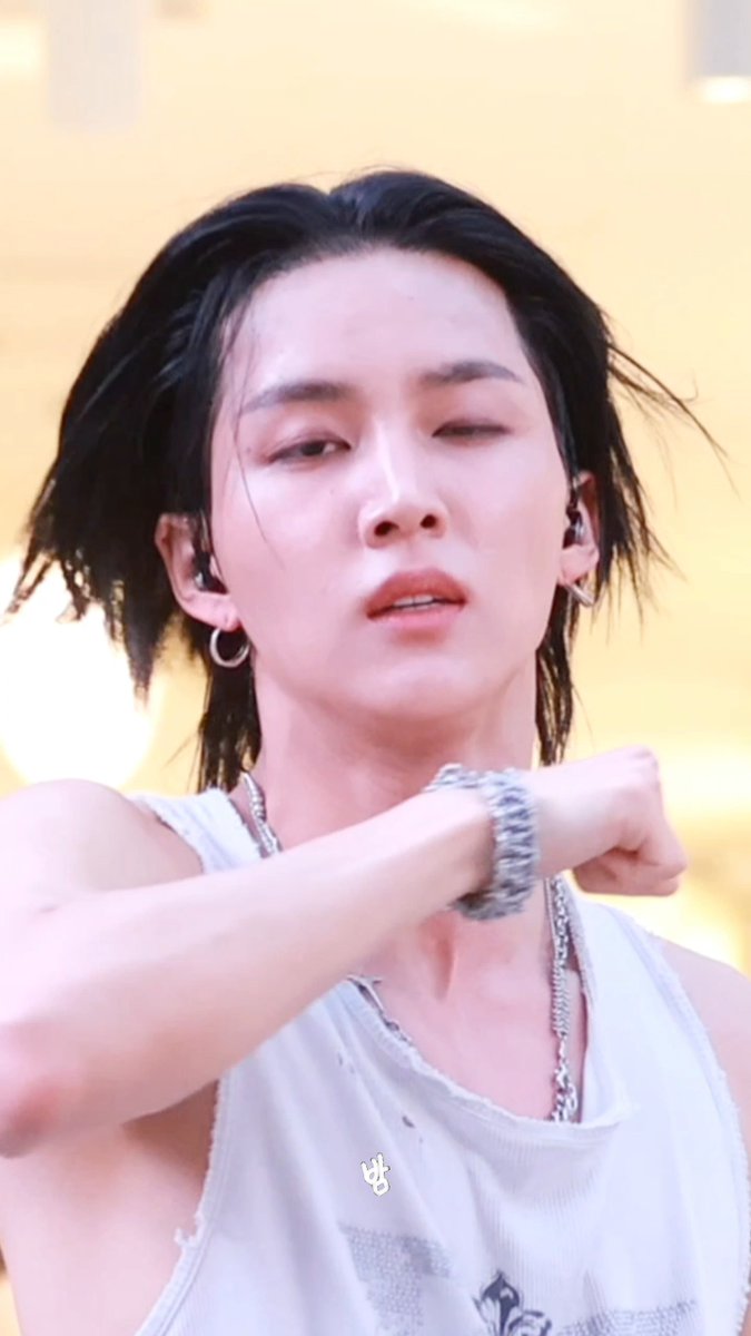 A close-up of a performer with wet dark hair wearing a distressed white tank top, silver chain necklace, and hoop earrings, with a Korean text sticker.