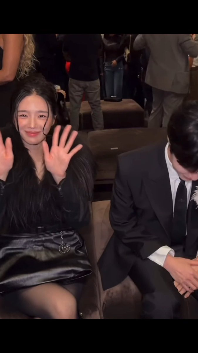 A woman with long black hair waves at the camera while seated next to a man in a black suit who is looking down, at what appears to be a formal event.