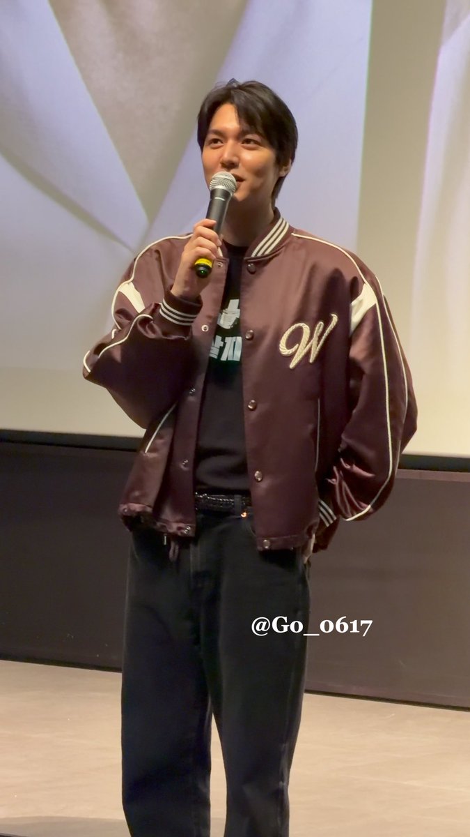 A young man speaks into a microphone on stage, wearing a maroon varsity jacket with a "W" embroidered on it over a black graphic t-shirt, with the watermark "@Go_0617."