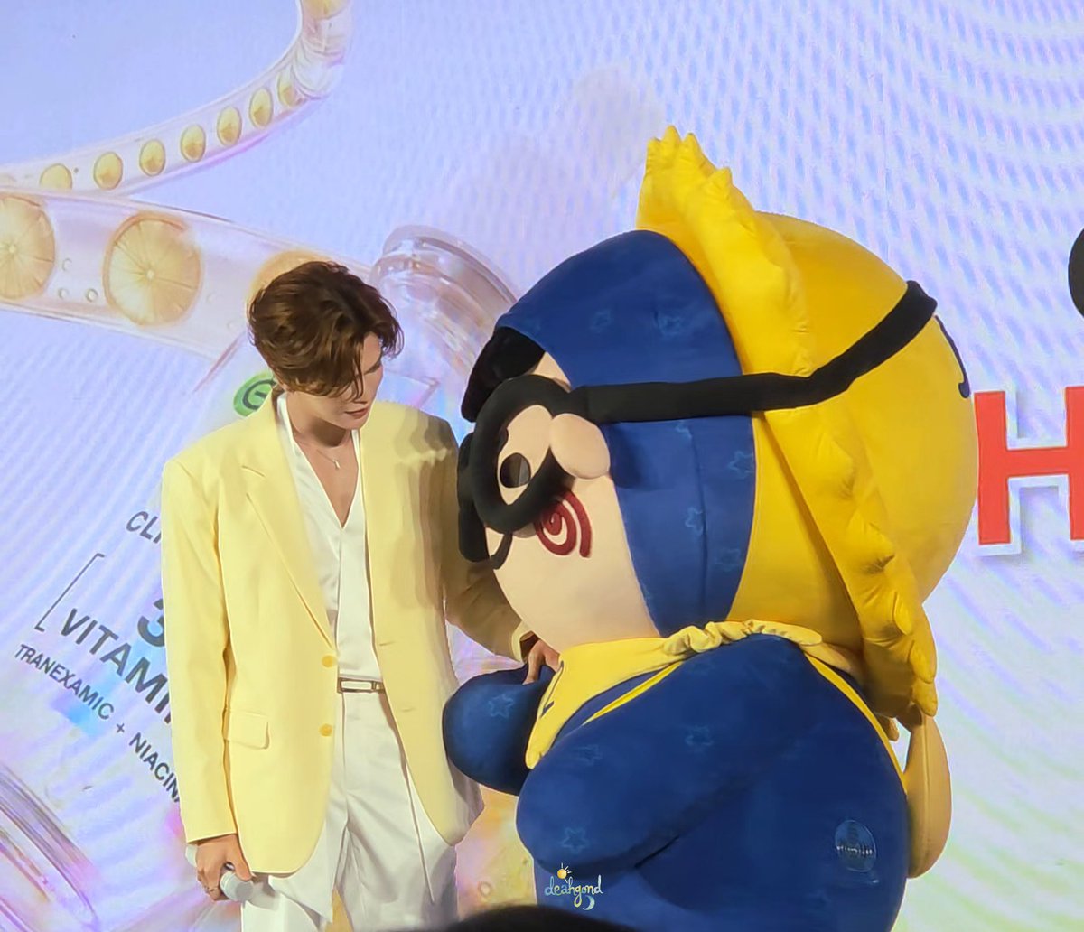 A young man in a pale yellow blazer stands beside a blue-and-yellow bird mascot (Jummo) with spiral red cheeks, posing in front of a Garnier Clinical Lab backdrop displaying "VITAMIN C" and "TRANEXAMIC + NIACINAMIDE." The image aligns with the post's caption about Mark and Jummo appearing together at the Garnier Clinical Lab event, with the hashtag #GarnierClinicalLabxMarkJummo visible in the watermark.