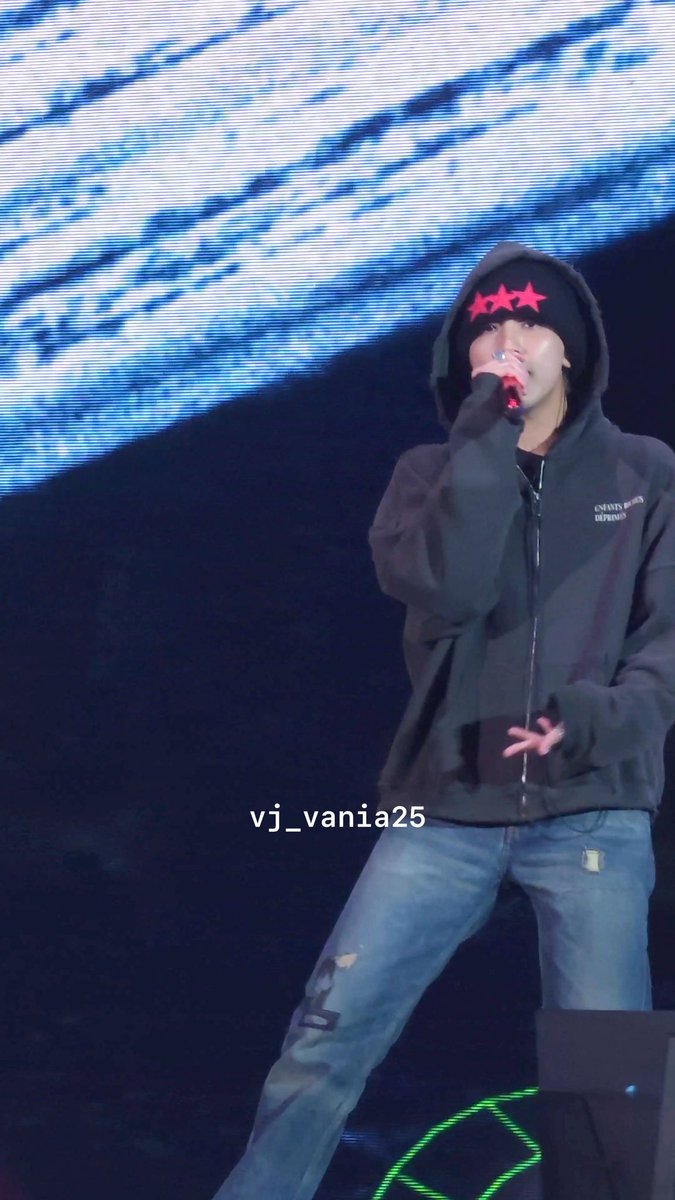A performer wearing a black hooded jacket, beanie with red stars, and ripped jeans sings into a microphone on stage, with a 'vj_vania25' watermark.