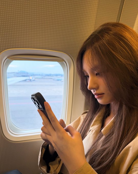 A young woman with long brown hair sits by an airplane window, focused on her smartphone as she travels.