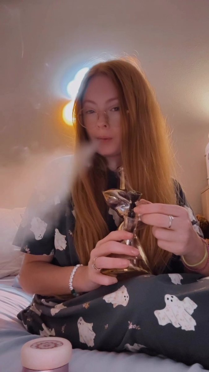 A woman with long reddish-blonde hair wearing ghost-patterned pajamas sits on a bed and smokes from a glass pipe, exhaling smoke.