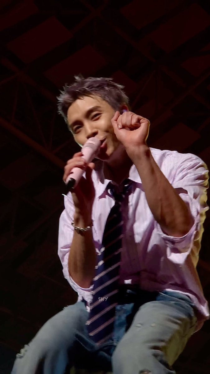 A young man with light purple-tinted hair performing on stage, singing into a pink microphone while wearing a white shirt and striped tie.