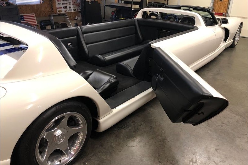 car_kobanashi's tweet photo. ダッジ バイパー RT/10 リムジン（1996 アメリカ）
1996年型「ダッジ バイパー RT/10」をベースに製作されたこのストレッチリムジンは、劇中車制作などで知られる米ラスベガスの「ユニーク・ムービー・カーズ」が手掛けた特装モデルである。大排気量アメリカンスポーツのシャシーを大胆に延長し、全長は25フィート（約7.6メートル）に達する。屋根のないオープン構造を保ちつつ、前席後方のロールバー造形を後端シートの後ろにも増設した独特のスタイリングを持つ。

車体中央の延長部分には対面式のロングベンチシートを新設。前席2名、車体後端の独立バケットシート2名と合わせ、販売時には最大12名乗車が可能と宣伝された。ホワイトのボディにブルーのストライプという伝統のカラーリングを纏い、荷室には大音量オーディオシステムを駆動するパワーアンプやオプティマ製バッテリーが搭載されている。

注目すべき変更点は動力部に存在する。本来の8.0リッターV型10気筒と6速MTは降ろされ、クライスラー製360立方インチ（約5.9リッター）「マグナム」V型8気筒エンジンへと換装。さらに変速機も純正設定のないオートマチック（AT）へと変更されているが、運転席にはダミーの6速MT用シフトノブが残され、視覚的な雰囲気を演出している。

この換装は、過激なV10よりも低速巡航やパレード運行における扱いやすさと熱管理を優先した現実的な選択といえる。獰猛な本格スポーツと華美なリムジン文化が融合した、極めて特異なアメリカンカスタムらしい車である。#車小噺