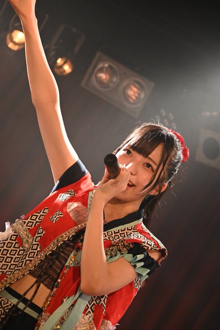 A young female performer in a red and patterned outfit sings into a microphone on stage, her arm raised under warm stage lighting.