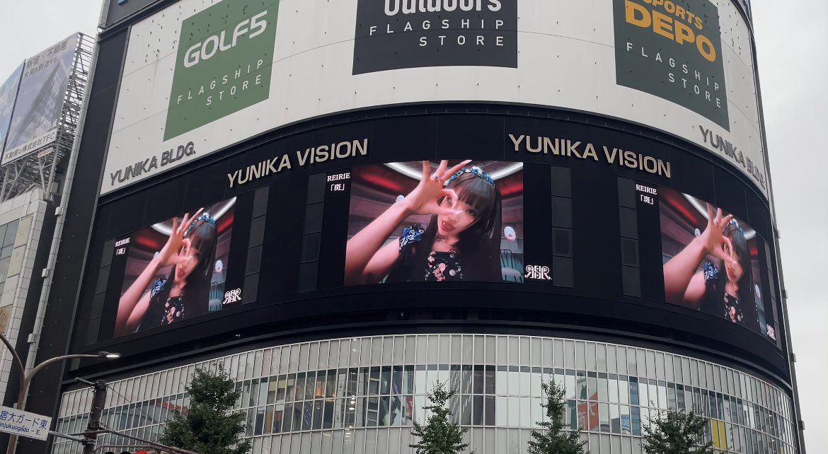 A street-level view of the YUNIKA VISION building in Japan featuring large curved digital billboards displaying a performer, alongside advertisements for 'GOLF5' and 'SPORTS DEPO' flagship stores.