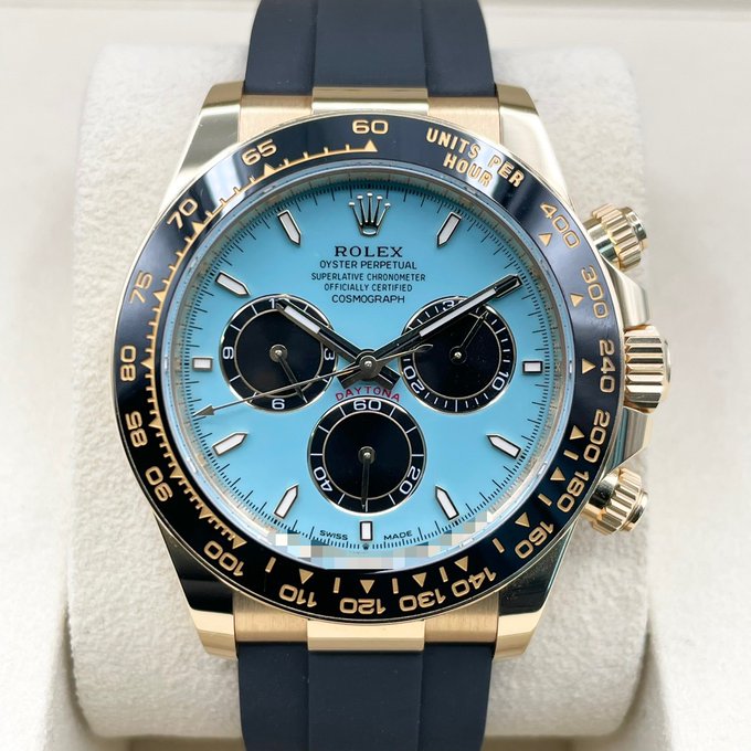 A close-up photograph of a luxury Rolex Oyster Perpetual Cosmograph Daytona watch featuring a light blue dial, yellow gold case, and dark blue ceramic bezel on a navy rubber strap.