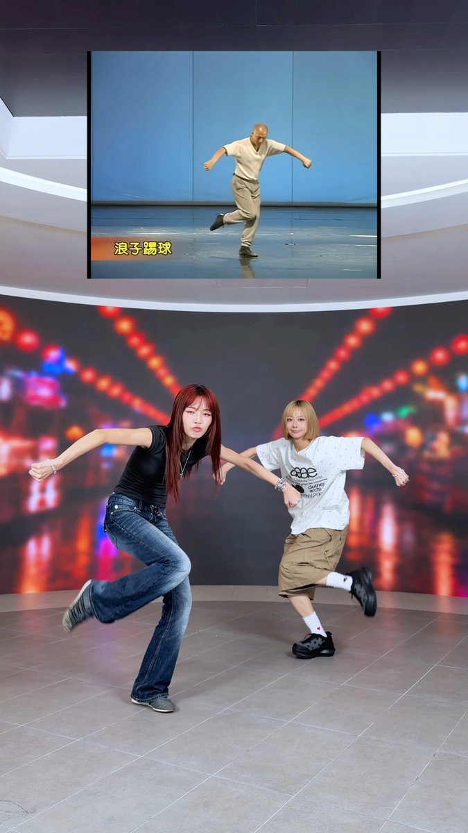 Two young women perform a synchronized dance move in a studio, with a screen behind them showing a bald man dancing with the Chinese text '浪子踢球'.