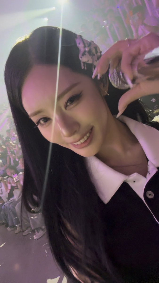 A close-up selfie of a smiling young woman with long dark hair, wearing a headband with cow-patterned charms, taken at what appears to be a concert venue.
