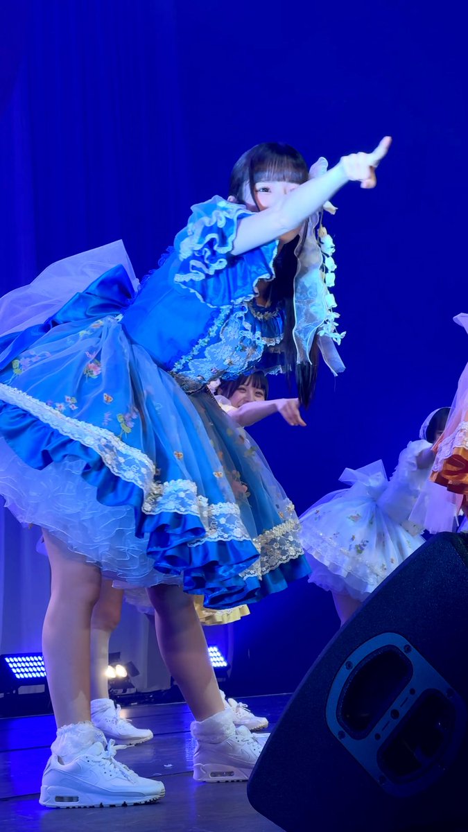 A young woman in an elaborate blue ruffled Lolita-style dress performs on stage, pointing forward under blue lighting.
