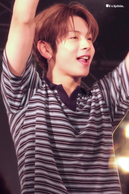 A young man with brown hair smiles joyfully with his arms raised, wearing a black and gray striped shirt with a dark collar, photographed on stage.
