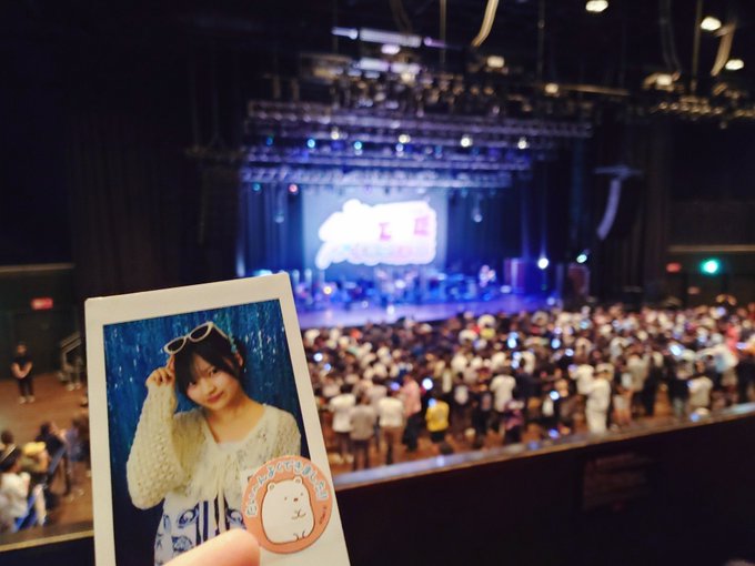 A polaroid photo of a young woman wearing sunglasses on her head is held up against the backdrop of a concert venue filled with a large crowd and a lit stage.