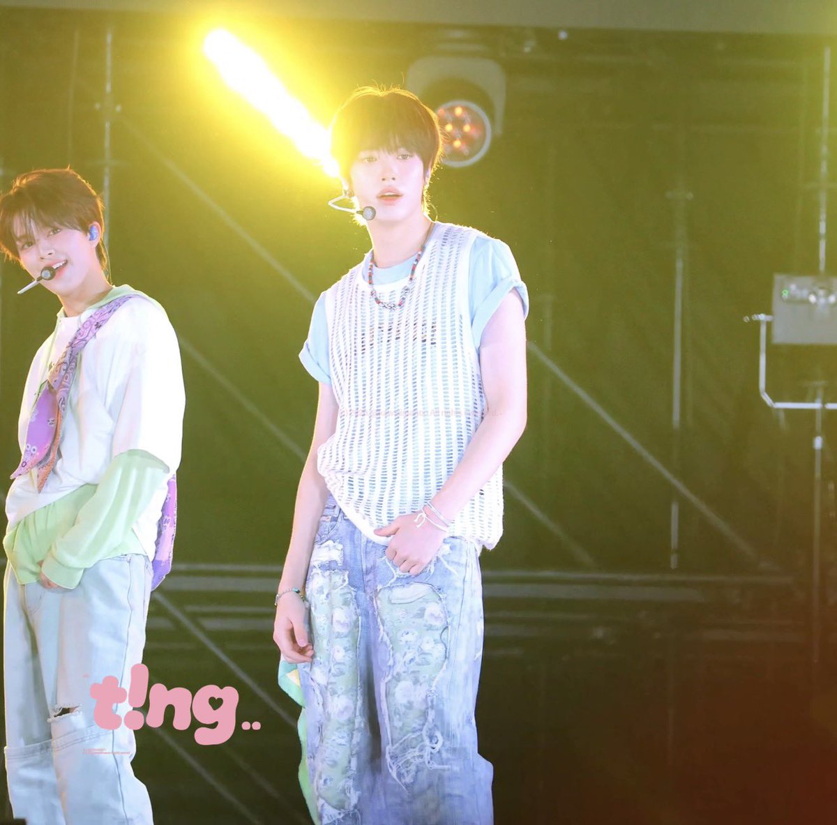 Two young male K-pop performers perform on stage with a bright spotlight behind them; one wears a white knit vest over a blue shirt with distressed jeans, with a 't!ng..' watermark visible.