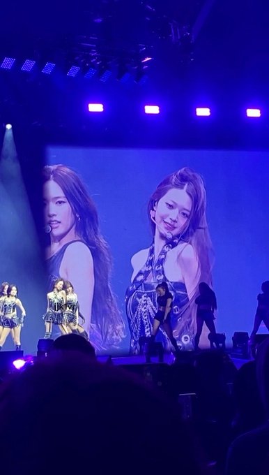 A concert scene shows performers on stage in blue lighting, with a large screen in the background displaying close-up images of two female artists.