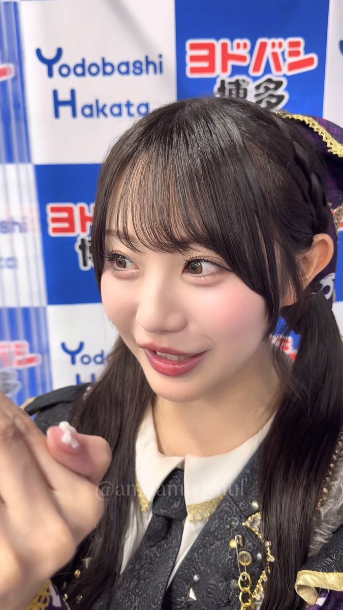 A young woman with dark hair styled in pigtails wearing an ornate black and white outfit, photographed in front of a blue and white 'Yodobashi Hakata' and 'ヨドバシ' branded backdrop.