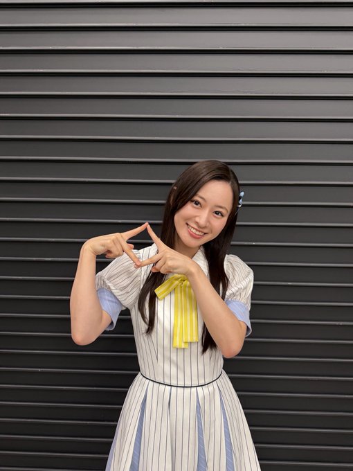 A smiling young woman with long dark hair, wearing a striped dress with a yellow bow, makes a triangle shape with her hands against a dark gray paneled wall.