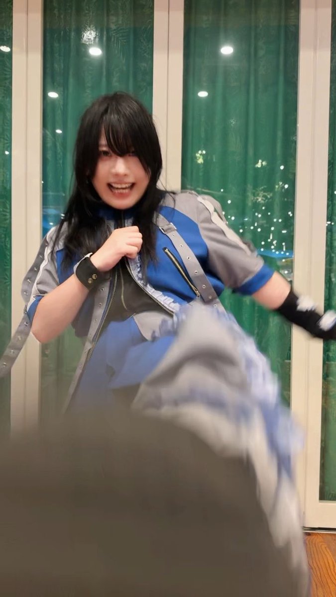 A young woman with dark hair wearing a blue and grey cosplay costume with black gloves, appearing to be in an action pose indoors.