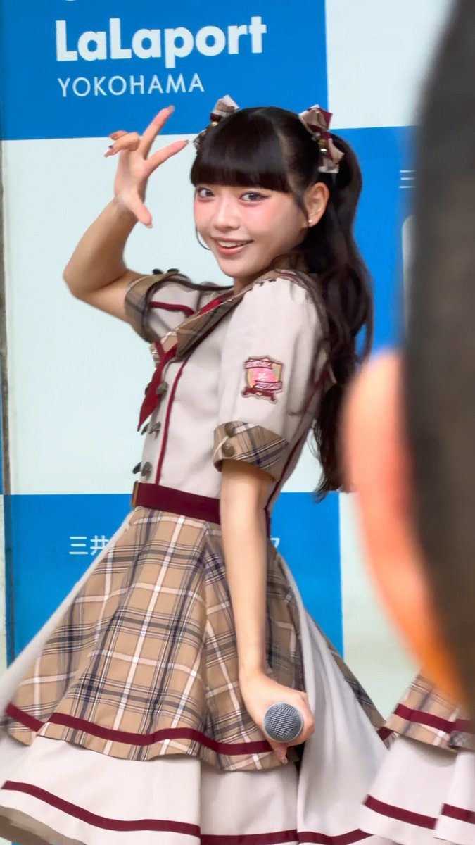 A young woman with dark hair in pigtails wearing a plaid idol-style dress poses with one hand raised, standing in front of a blue 'LaLaport YOKOHAMA' sign.