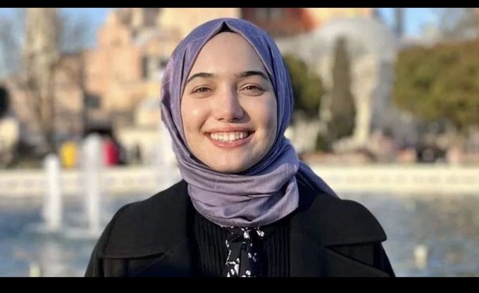 A smiling young woman wearing a purple hijab and a black coat, posing outdoors with a fountain visible in the background.
