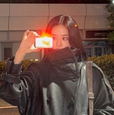 A young woman taking a mirror selfie outdoors at night, holding up a camera with a bright flash, wearing a black leather jacket.