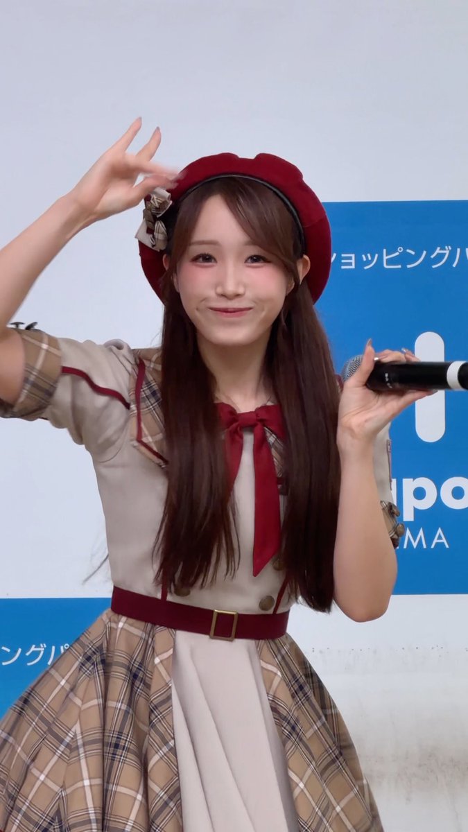 A young woman, likely a Japanese idol, performing on stage while wearing a red beret, beige and plaid dress with a red bow, and holding a microphone.