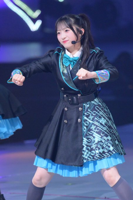 A young woman with long dark hair performs on stage wearing a black and teal costume with a bow tie, a pleated skirt with musical note patterns, and a headset microphone.