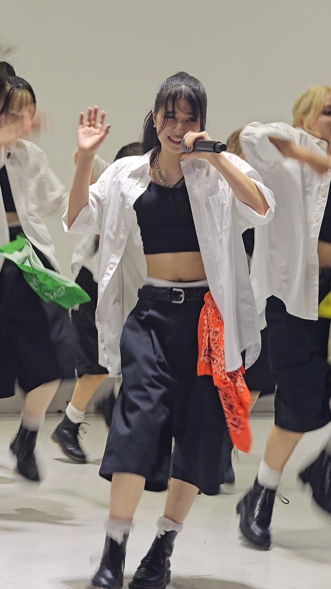 A young woman singing into a microphone while performing on stage, wearing a white open shirt over a black top and black shorts with an orange bandana.
