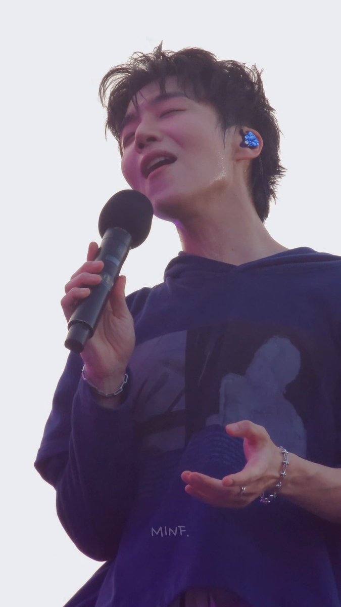 A male performer with messy dark hair sings passionately into a microphone on stage, wearing a dark oversized shirt and a blue in-ear monitor, with 'MINF.' visible at the bottom.