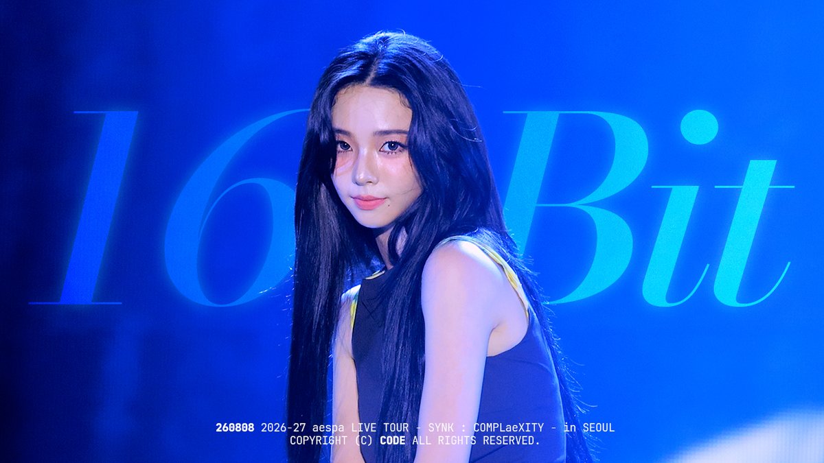 A young woman with long dark hair poses in front of a blue backdrop displaying the text '16 Bit'. Text at the bottom reads '260808 2026-27 aespa LIVE TOUR - SYNK : COMPLæXITY - in SEOUL' and 'COPYRIGHT (C) CODE ALL RIGHTS RESERVED.'