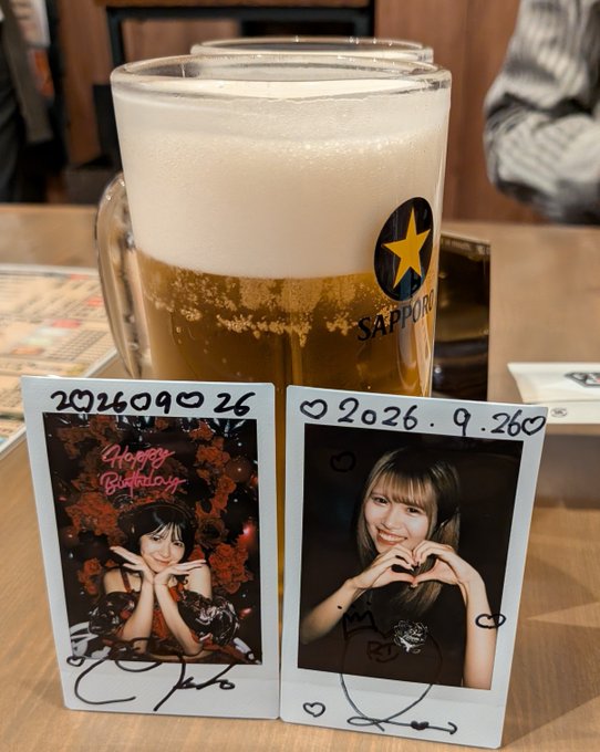A mug of Sapporo beer sits on a table behind two polaroid photos dated 2026.9.26, one showing a woman with 'Happy Birthday' written on it.