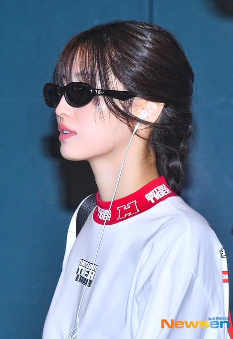 A side profile of a woman wearing black sunglasses, white earphones, and a white 'RUNNING MAN' shirt with a red collar, photographed against a blue background with a 'Newsen' watermark.